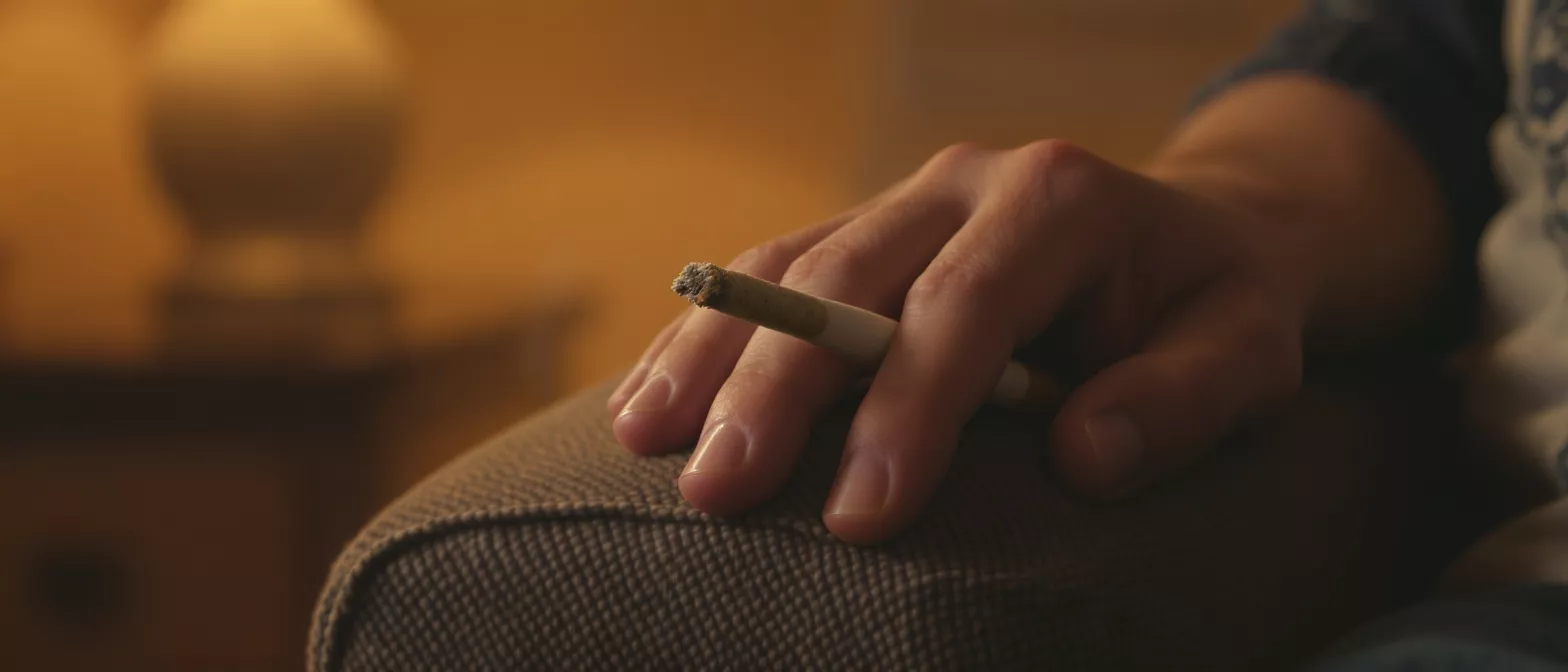 Close-up of a hand holding a lit cigarette with ash, resting on a textured surface.