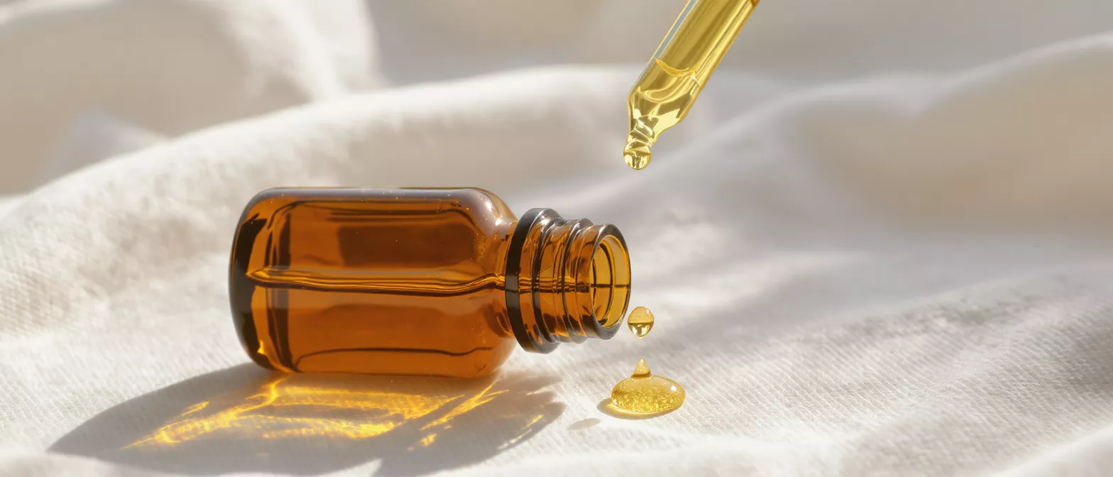 Amber glass bottle with a dropper dispensing golden oil onto a white textured fabric.