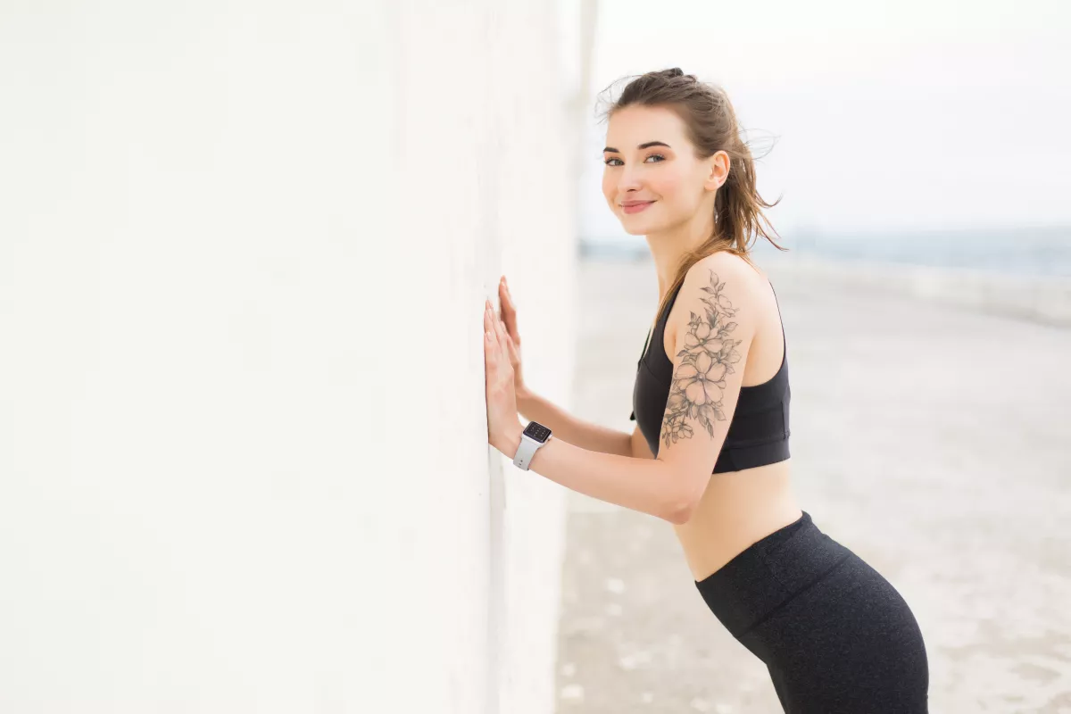 Fit woman in activewear stretches by beach, displaying arm tattoo and smartwatch.