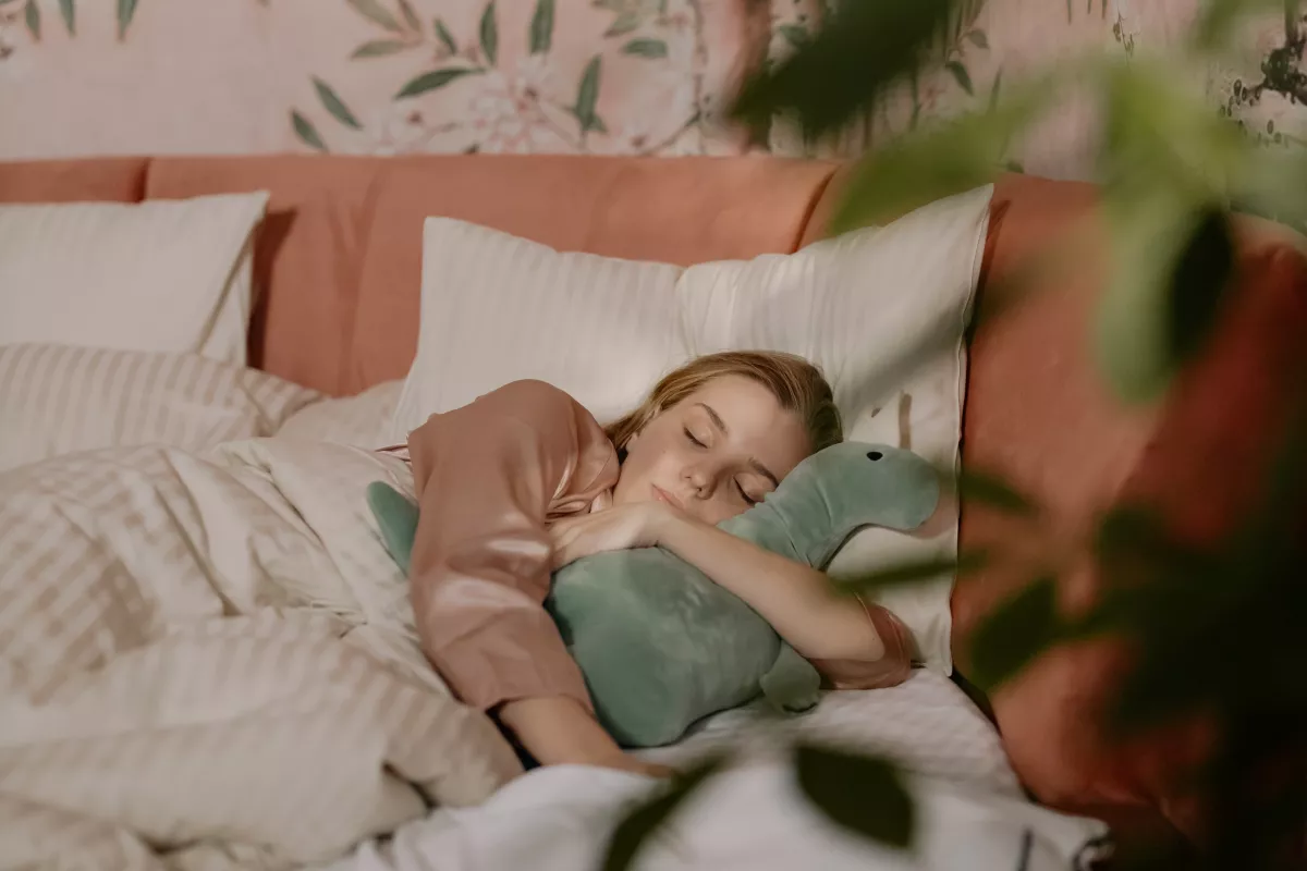 Person sleeping peacefully with dinosaur plushie in a serene, plant-filled bedroom.
