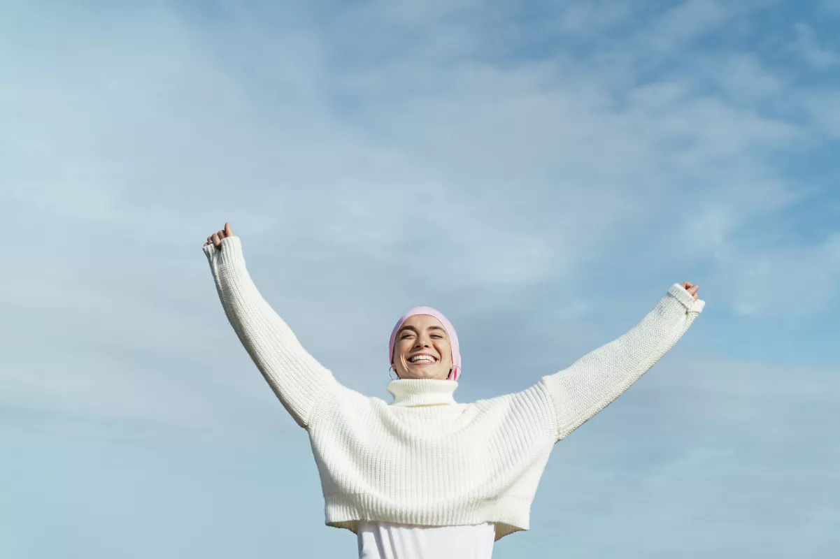 Person in headscarf and sweater celebrates with arms raised against a bright blue sky.