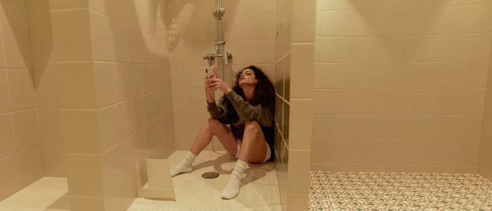 Woman Sitting in Shower with Phone A woman with curly hair sits on the floor of a tiled shower, holding a phone.