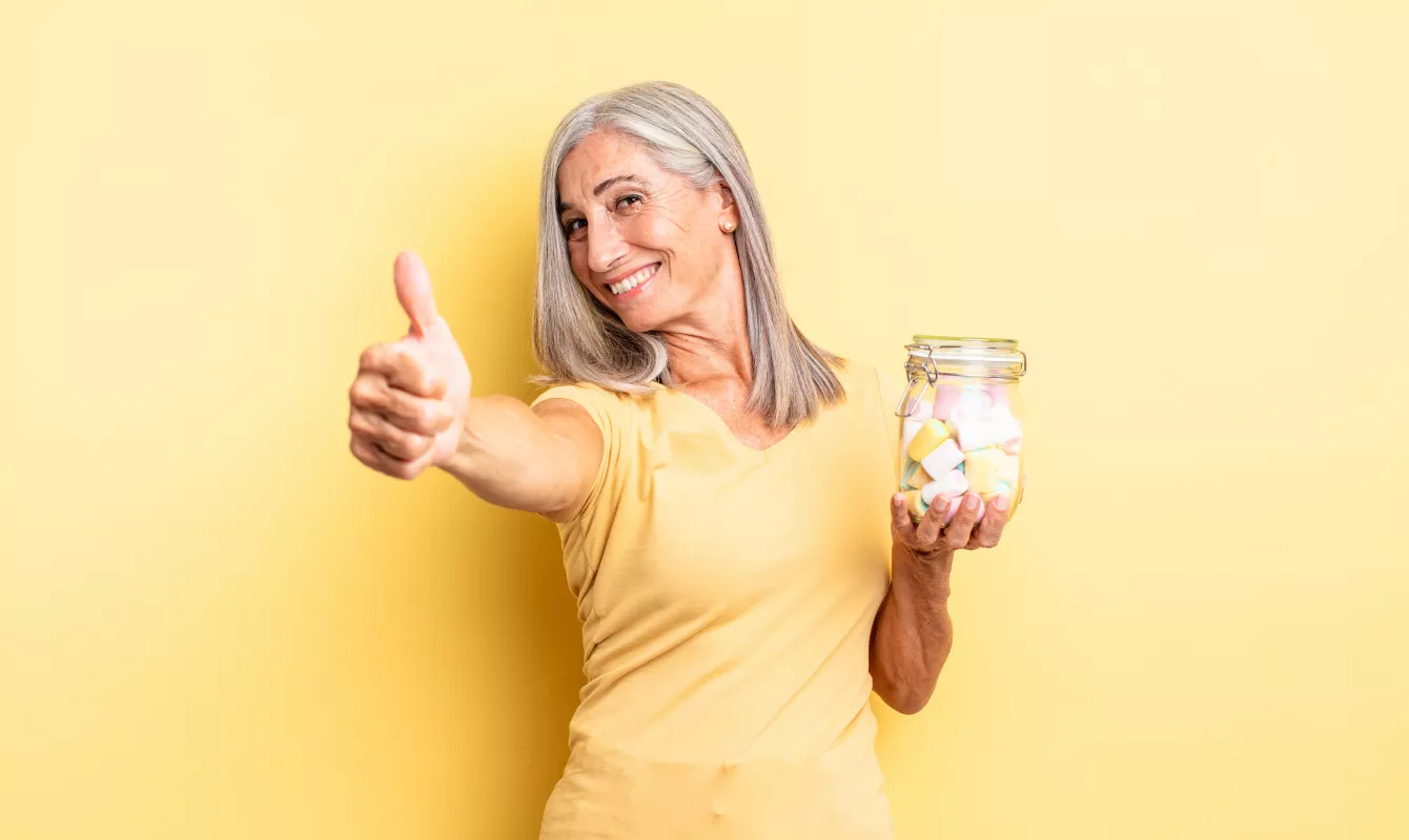 Senior woman with grey hair smiling, thumbs up, holding CBD marshmallow jar against yellow background.