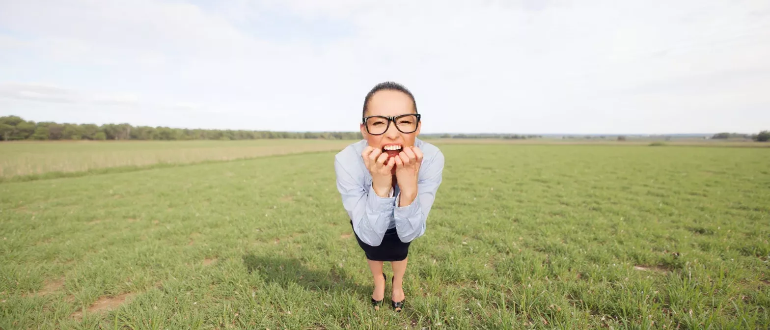 Woman in glasses looks stressed, biting nails in a wide-open field.