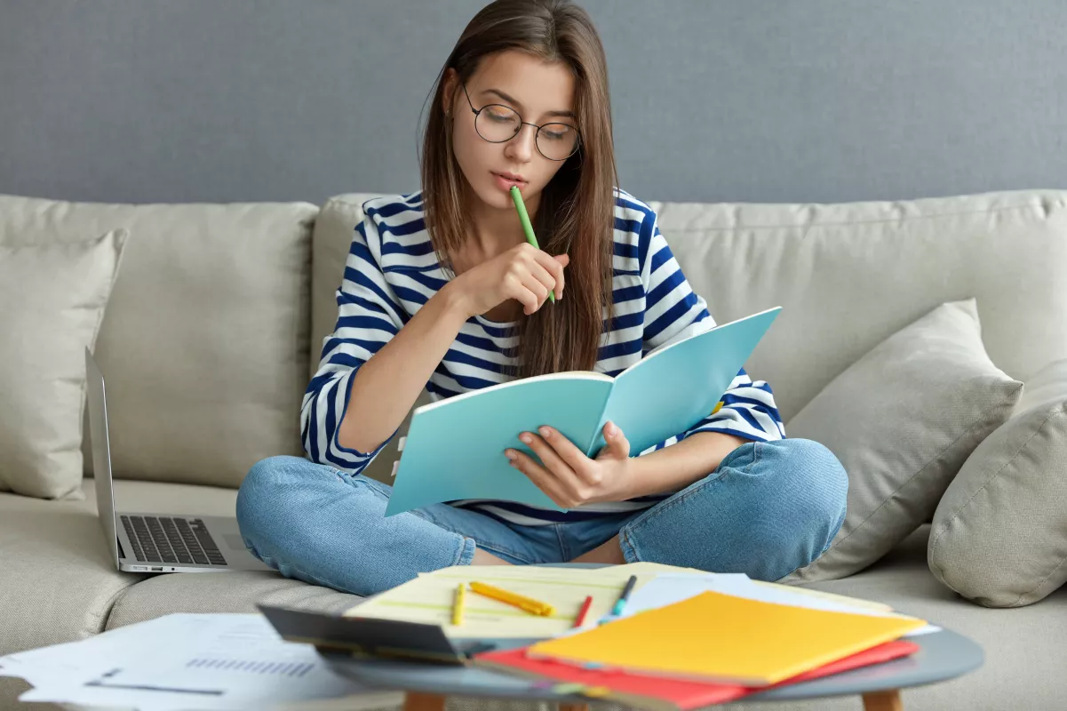 Woman studying at home with laptop and notes on couch.