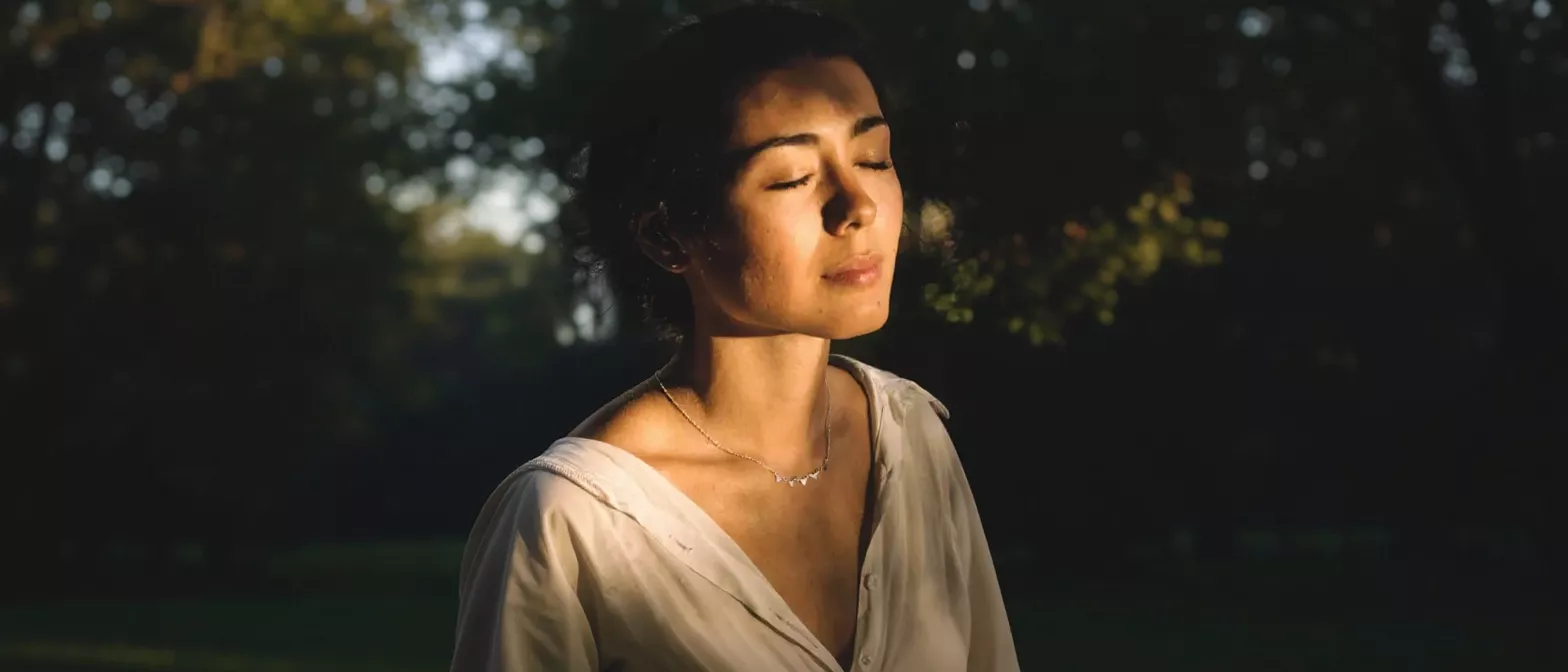 A woman with closed eyes, illuminated by sunlight, meditating outdoors.
