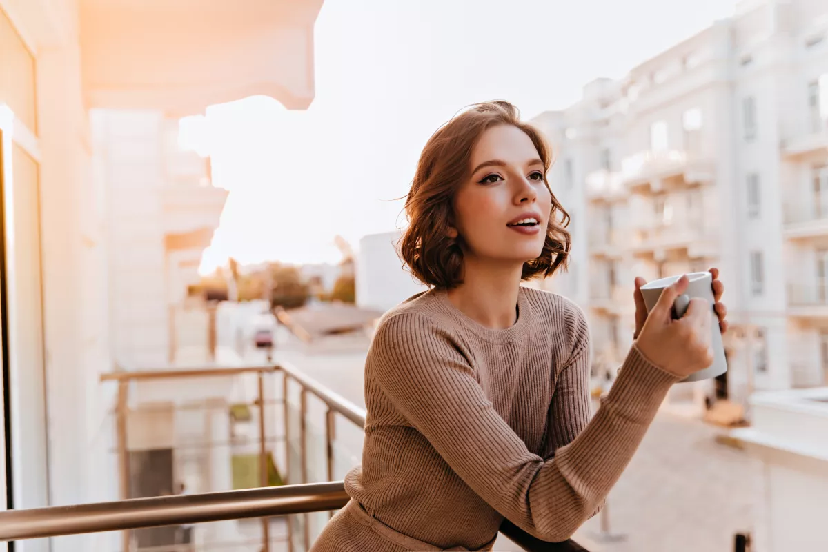 Woman enjoys coffee on balcony at dusk, peaceful urban setting.