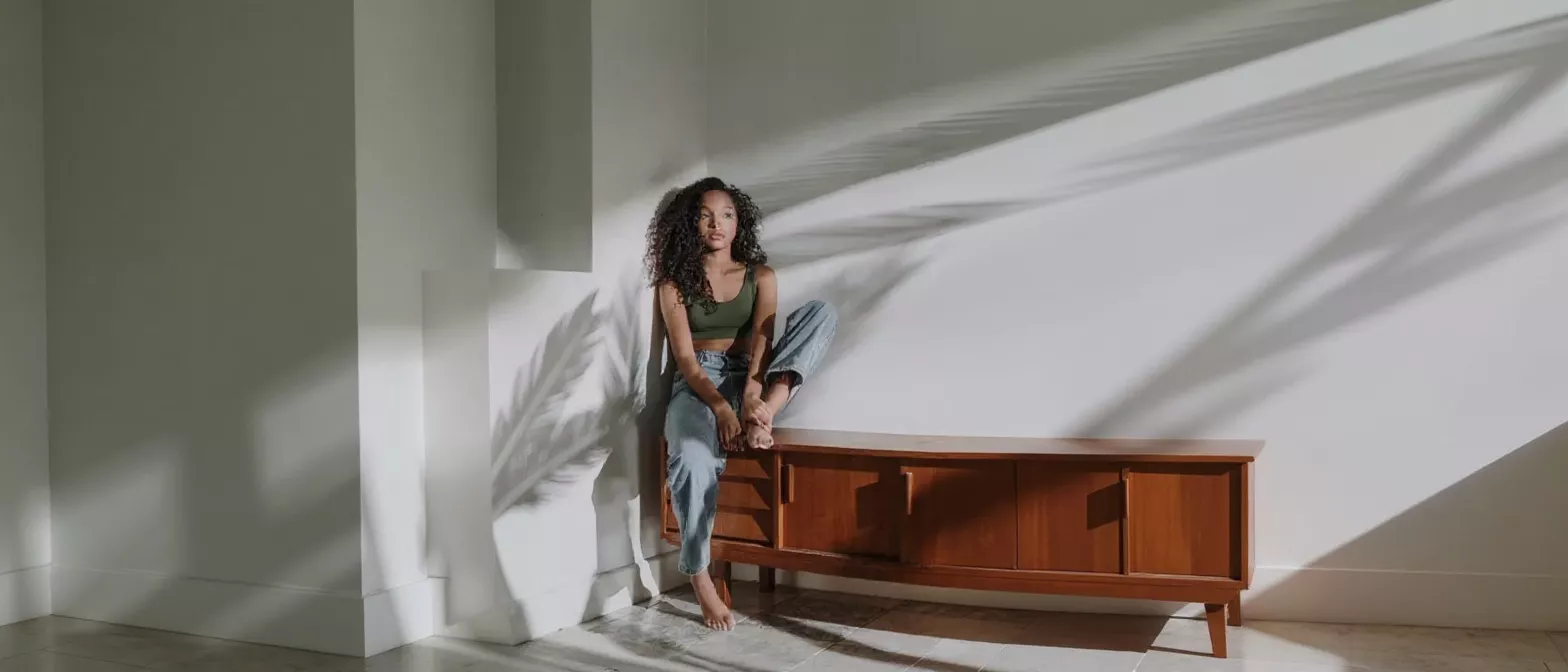 Young woman with curly hair sitting on a wooden cabinet, bathed in sunlight and shadows.