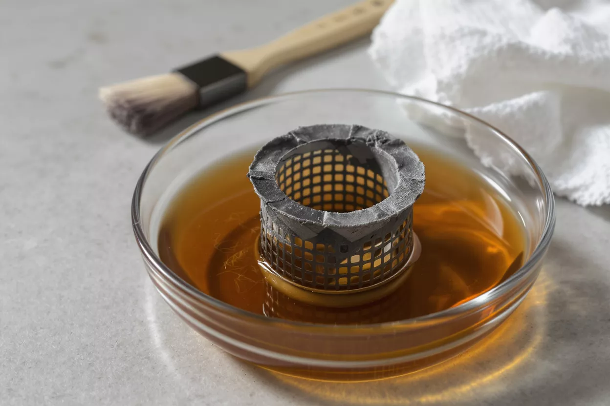 A dirty mesh appliance filter soaking in a glass bowl of amber-colored liquid.
