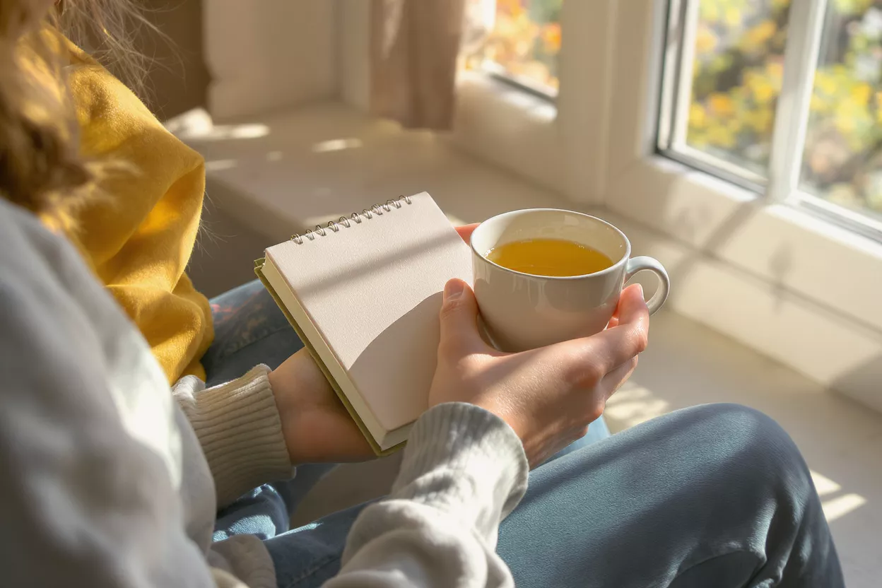 Person holding a journal and tea cup by a sunlit window.