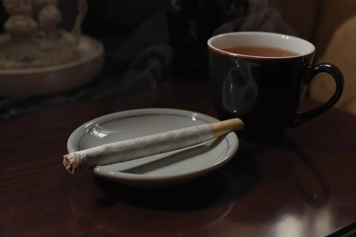 A lit joint on a small plate next to a steaming cup of tea.