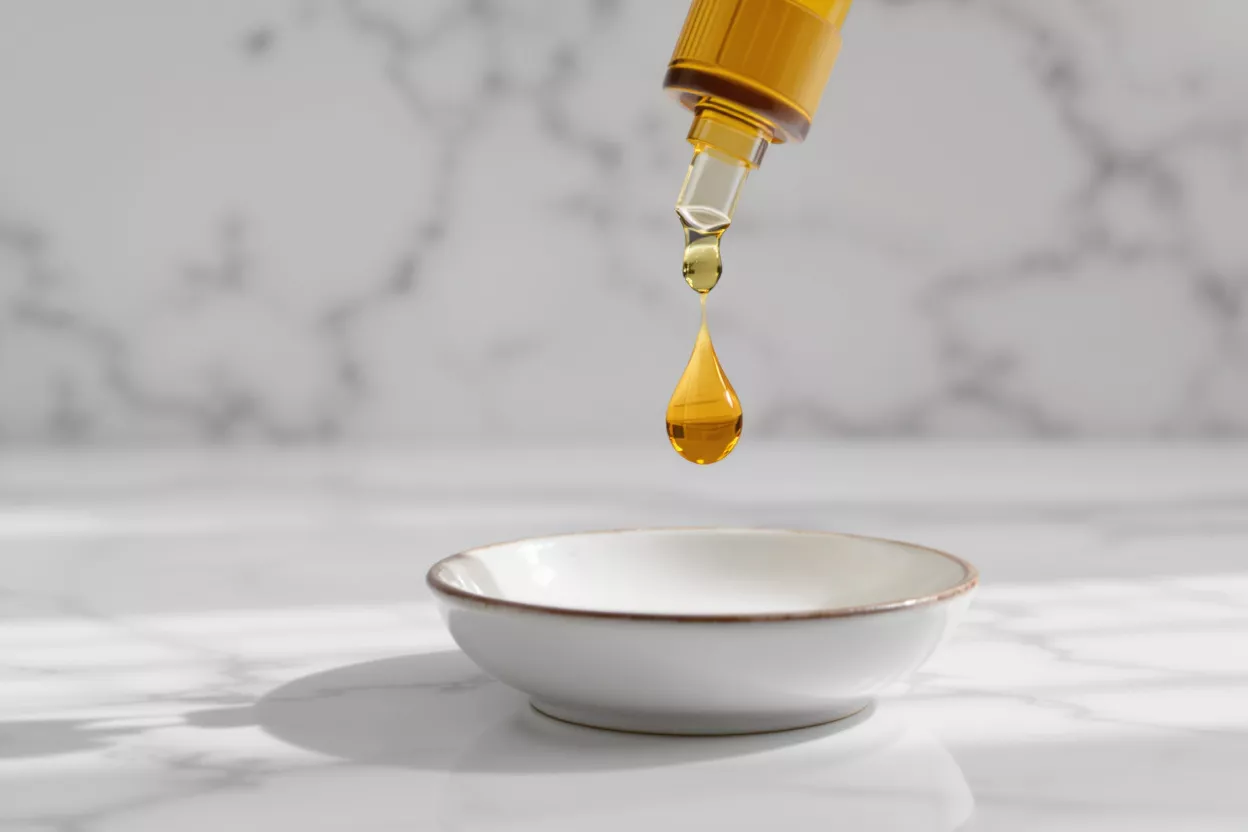 A golden oil drop falling from a dropper into a white bowl on a marble surface.