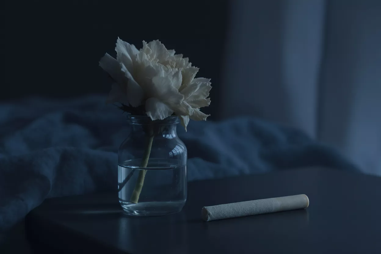 Flower Vase and Cigarette White flower in a clear vase next to a cigarette on a dark table.