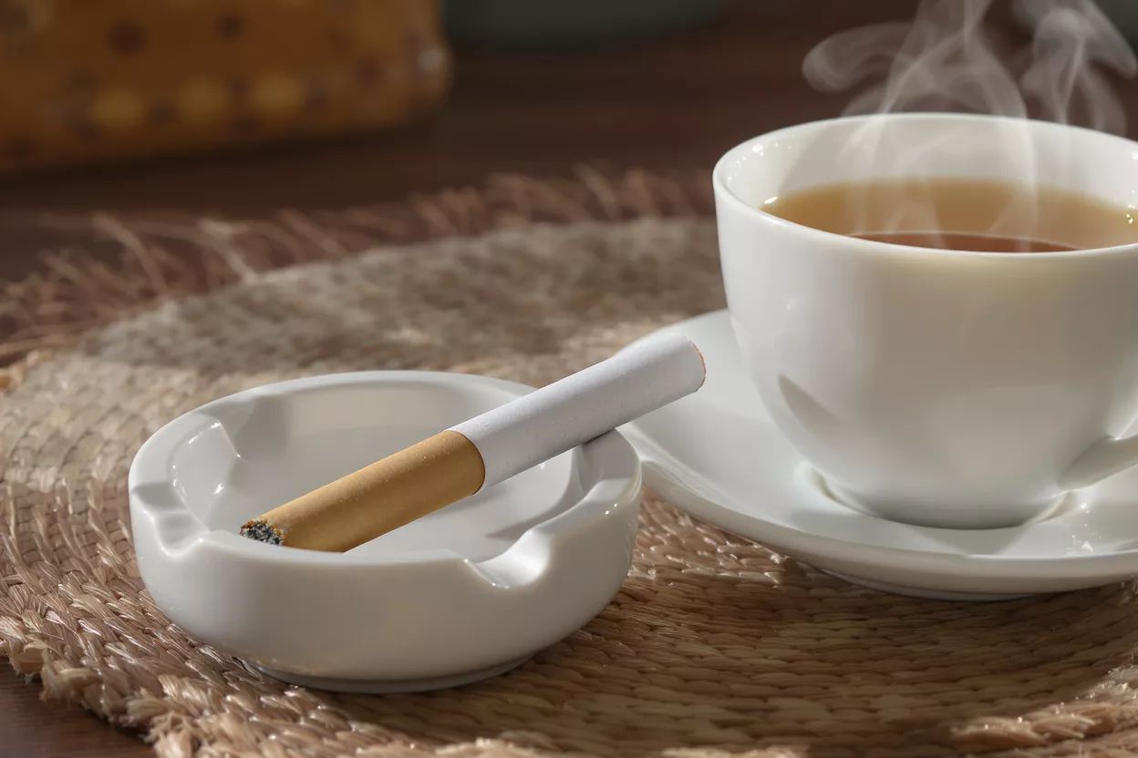 A lit cigarette in an ashtray next to a steaming cup of coffee.