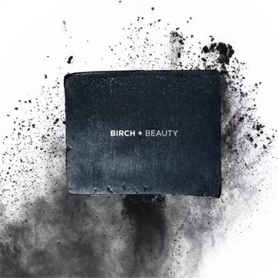 Birch + Beauty detoxifying charcoal soap with scattered particles on white background.