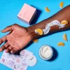 Skincare routine with cream, dark-skinned arm, mandarin segments, and colorful stickers on blue background.
