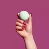 Hand holding green bath bomb with maroon nails on magenta background.