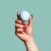Hand holding light blue bath bomb with magenta nails against turquoise background.