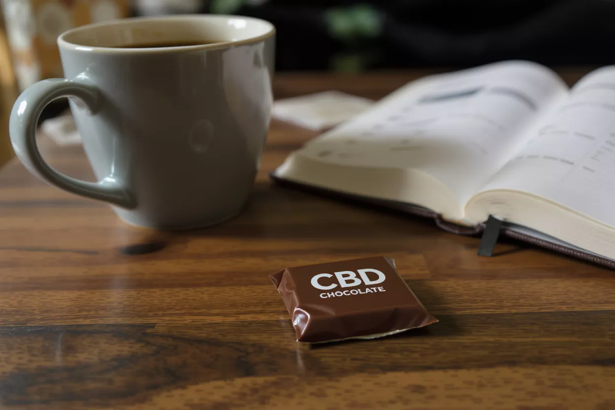 Una tableta de chocolate con CBD, una taza de café y un libro abierto sobre una mesa de madera.