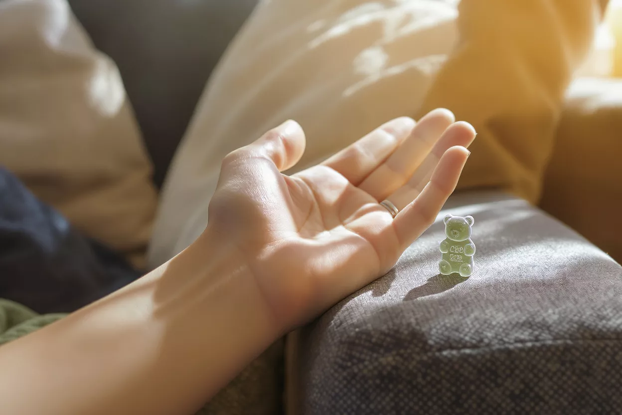 A person's hand resting beside a small green CBD gummy bear on a couch.
