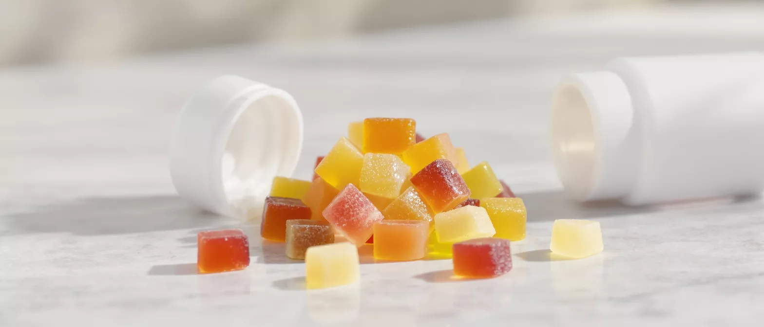 Gominolas cuadradas vibrantes de color amarillo, naranja y rojo derramadas de un frasco blanco.