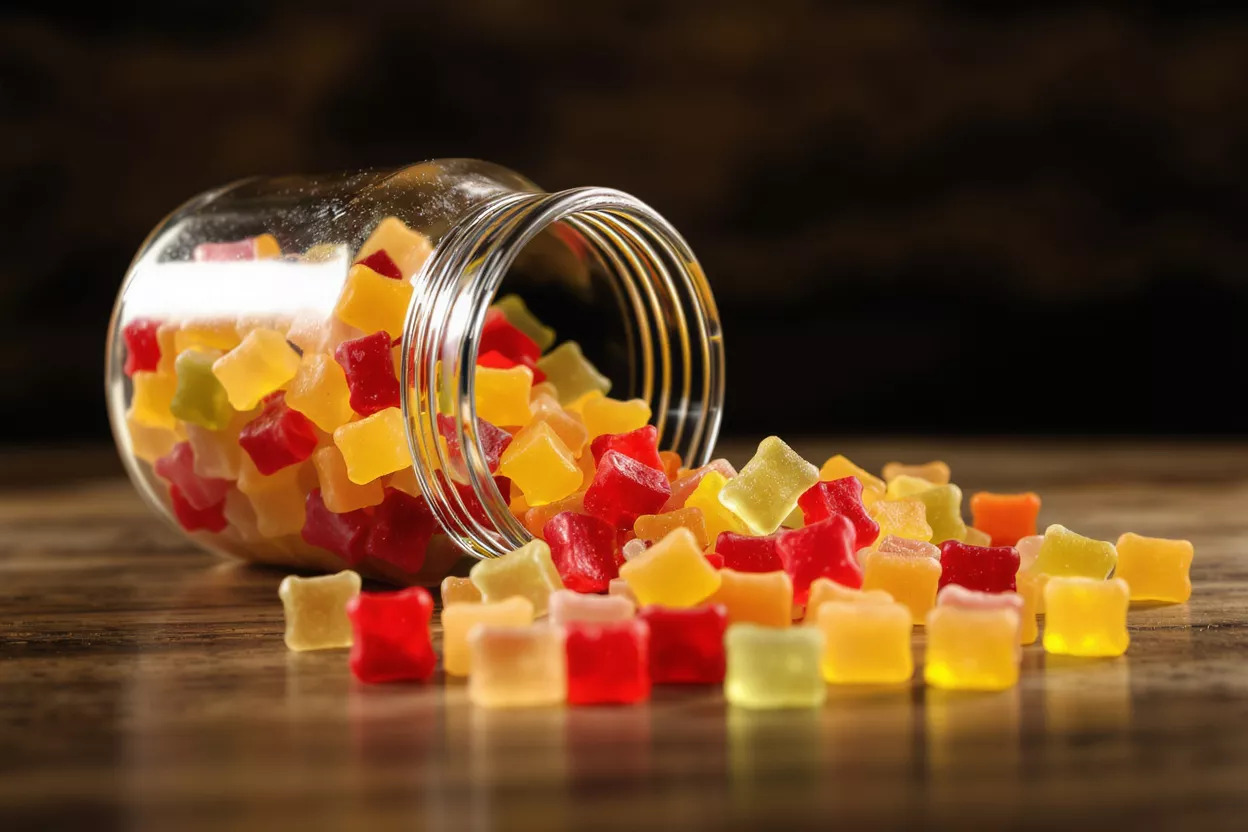 A glass jar spilling colorful square gummy candies onto a wooden surface.