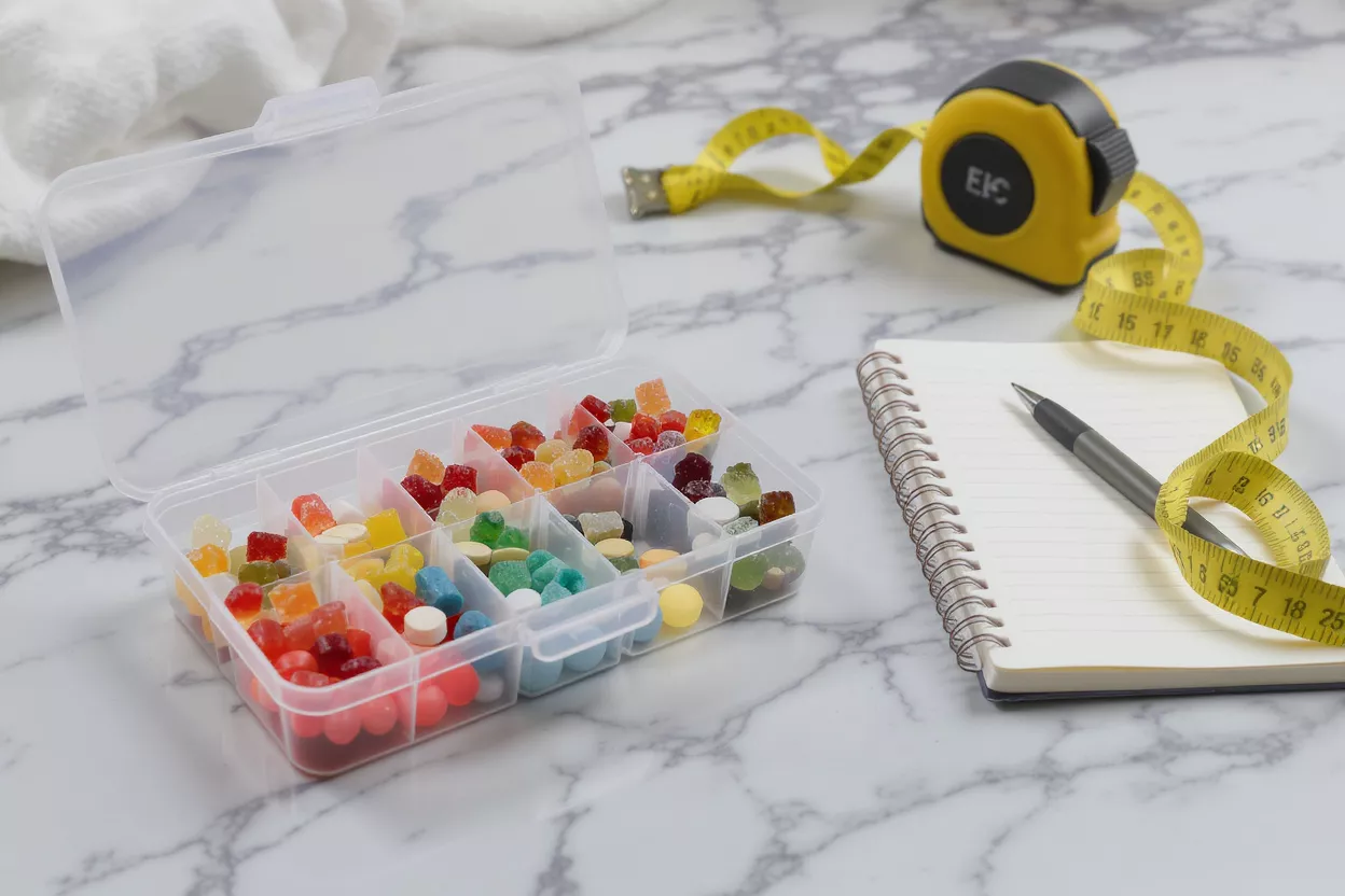 Boîte organisatrice transparente avec des bonbons gélifiés colorés, un ruban à mesurer, un carnet et un stylo sur du marbre.