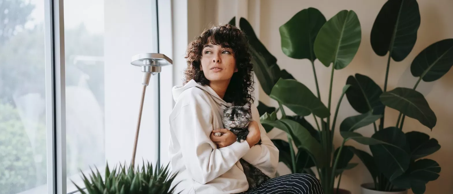 Young woman holding a cat, looking out a window with plants nearby.