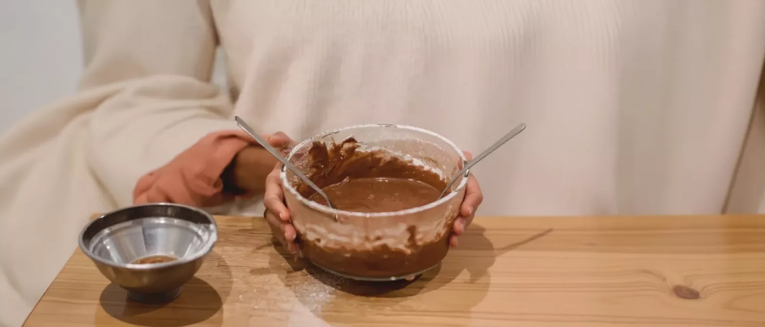 Hands holding a glass bowl of chocolate batter with spoons, next to a small metal bowl.