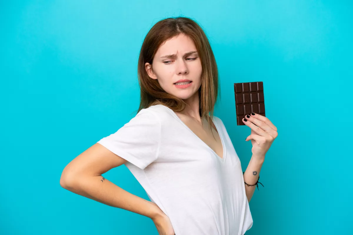 Young adult contemplating choice with chocolate bar against teal backdrop.