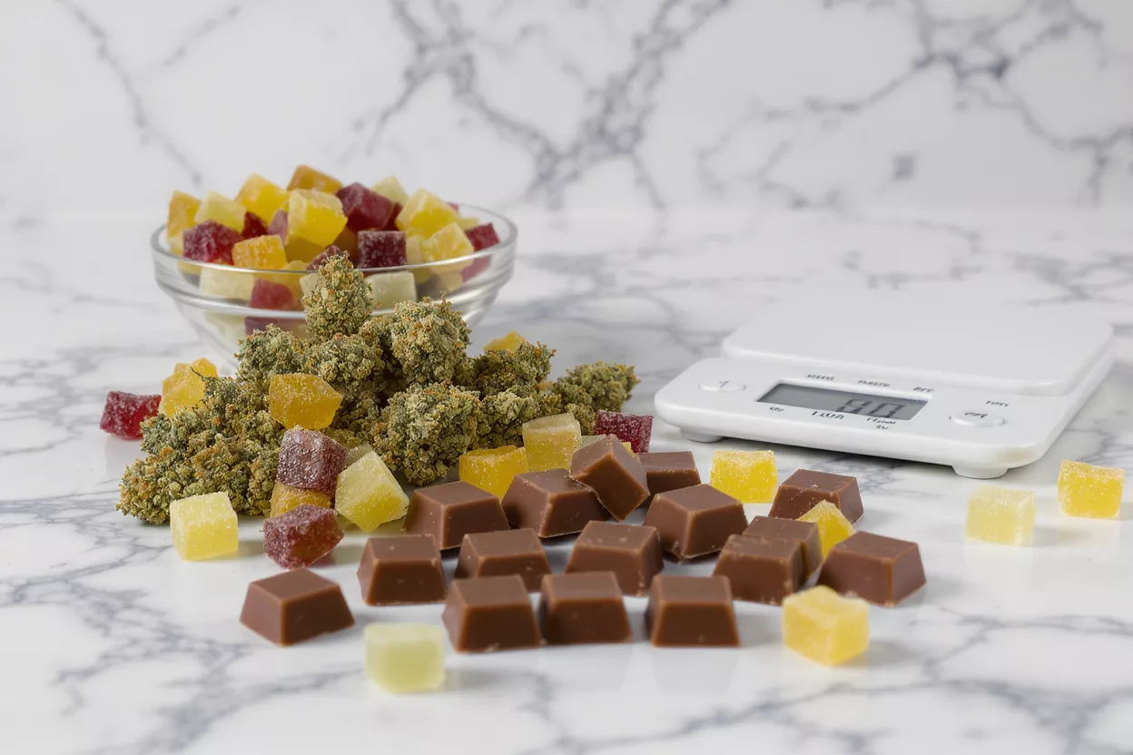 Cannabis flower, colorful gummies, chocolate edibles, and a digital scale on marble.