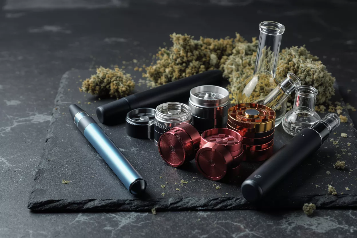 Various cannabis accessories including vapes, grinders, and glass beakers on a dark surface.