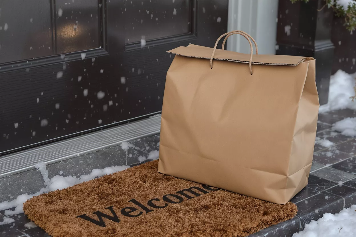 Delivery Bag on Snowy Welcome Mat Brown paper delivery bag on a 'Welcome' mat in snow by a dark door.
