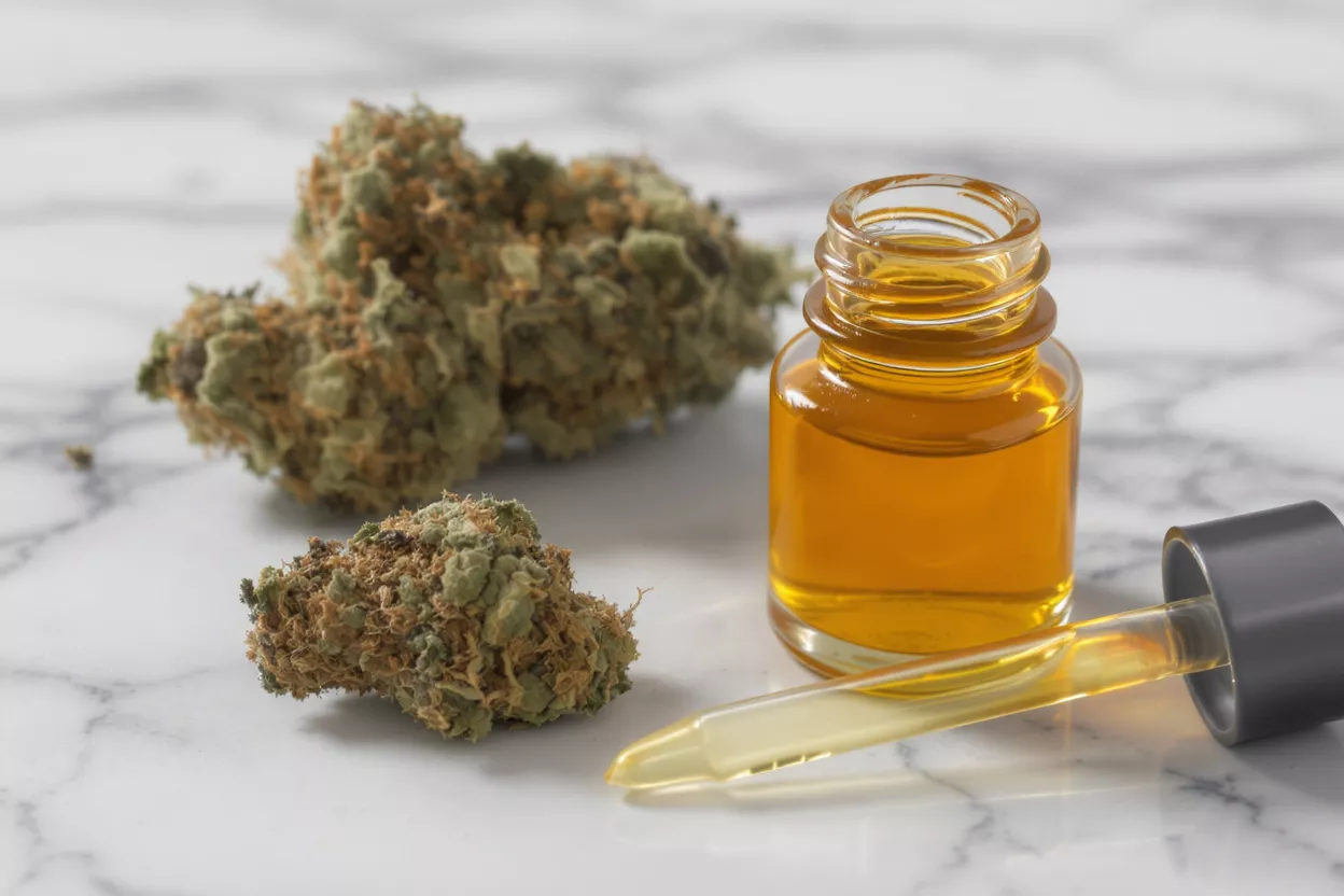 Cannabis buds and a bottle of CBD oil with a dropper on a marble surface.