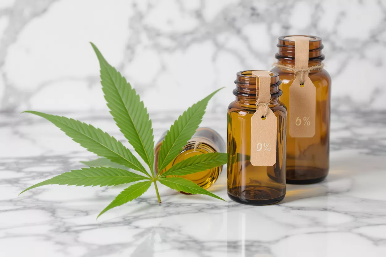 CBD Oil Bottles with Cannabis Leaf Cannabis leaf next to amber bottles with 0% and 6% labels on marble background.
