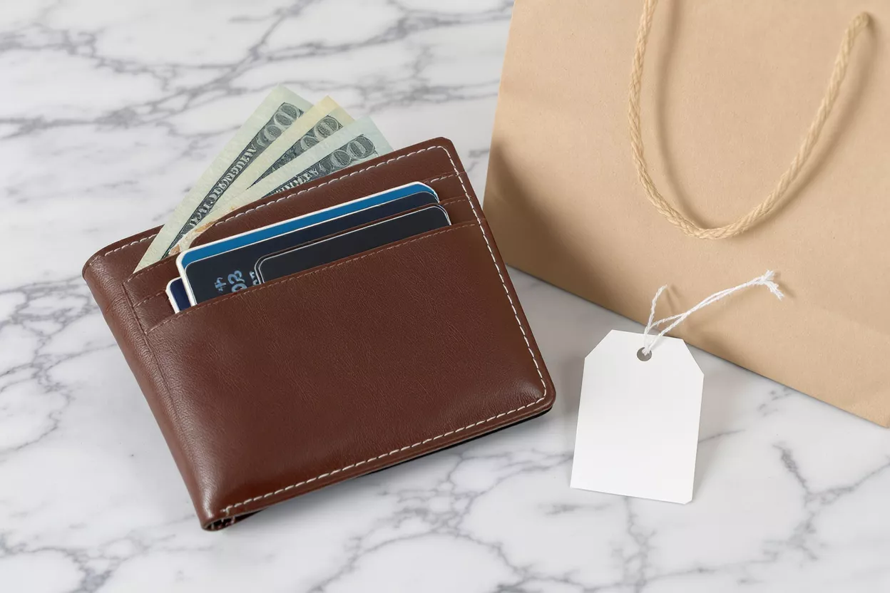 Wallet, Money, and Shopping Brown wallet with cash and cards, shopping bag, and blank tag on marble.