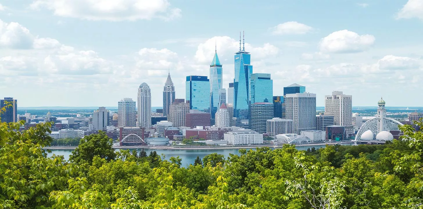 Modern city skyline with skyscrapers, lush greenery, and calm water reflecting a vibrant sky.