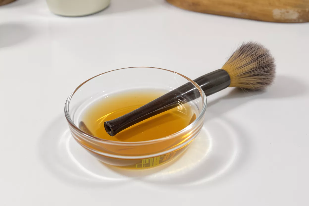 A pastry brush with a dark handle resting in a small glass bowl of oil.