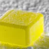 Close-up of a melting yellow ice cube against a blurred greyish-white background.