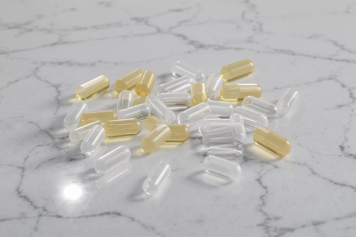 A mix of clear empty and yellow liquid-filled gel capsules on a marble surface.