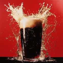 Dark beer overflowing in pint glass with splash, red background, and frothy foam.