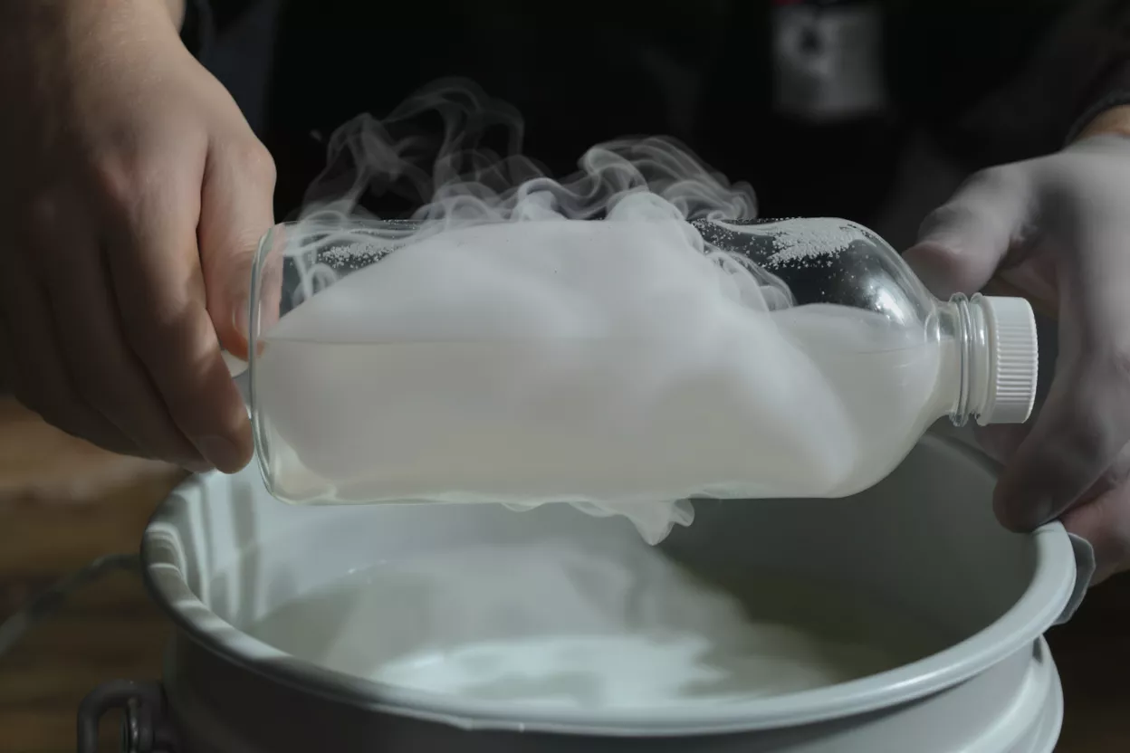 Gloved hands pouring white smoke from a clear plastic bottle into a grey bucket.