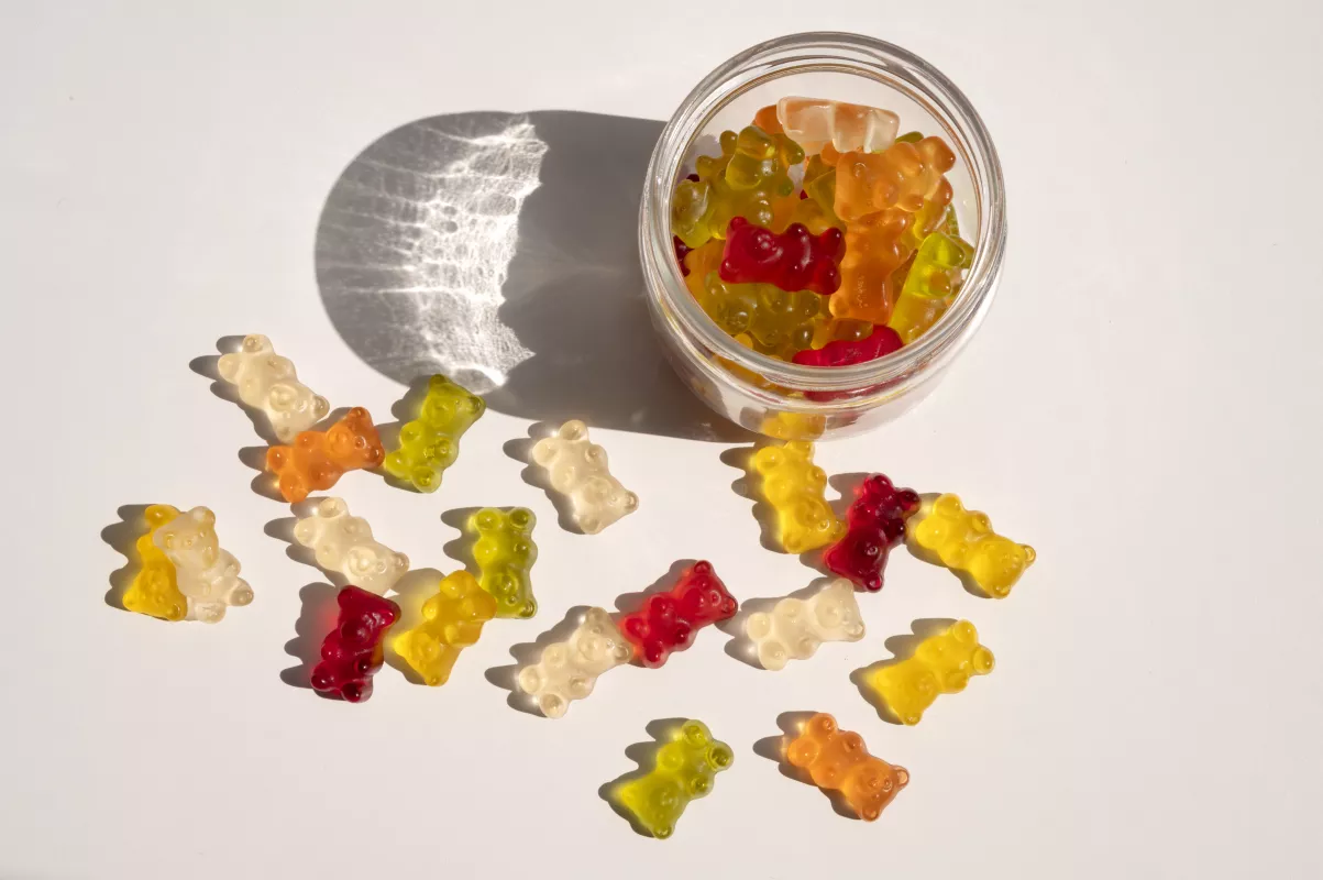 Colorful gummy bears tumble from jar on white surface with striking shadow.