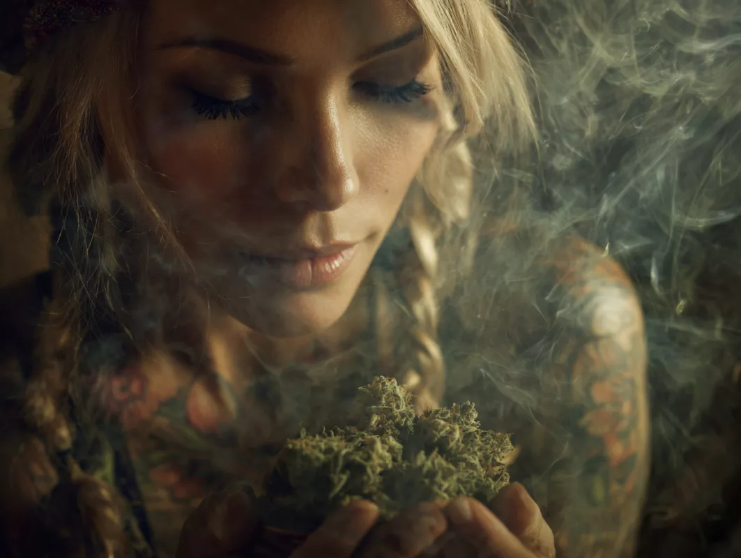 Serene woman meditating, holding cannabis, surrounded by smoke, showcasing bohemian style and tattoos.