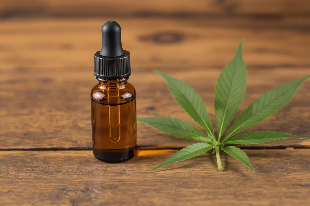 CBD Oil Bottle and Cannabis Leaf Amber dropper bottle of CBD oil next to a fresh green cannabis leaf on wood.