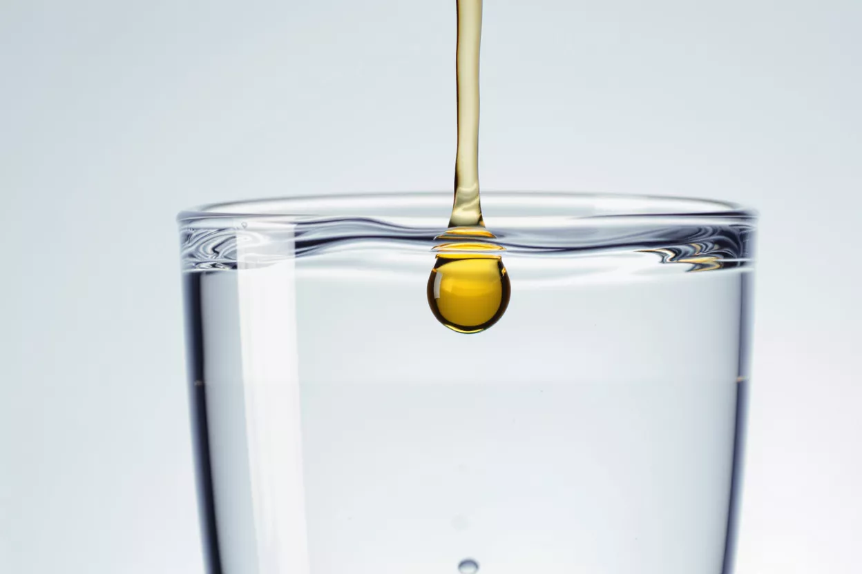 A golden oil drop detaching from a stream into clear water in a glass.