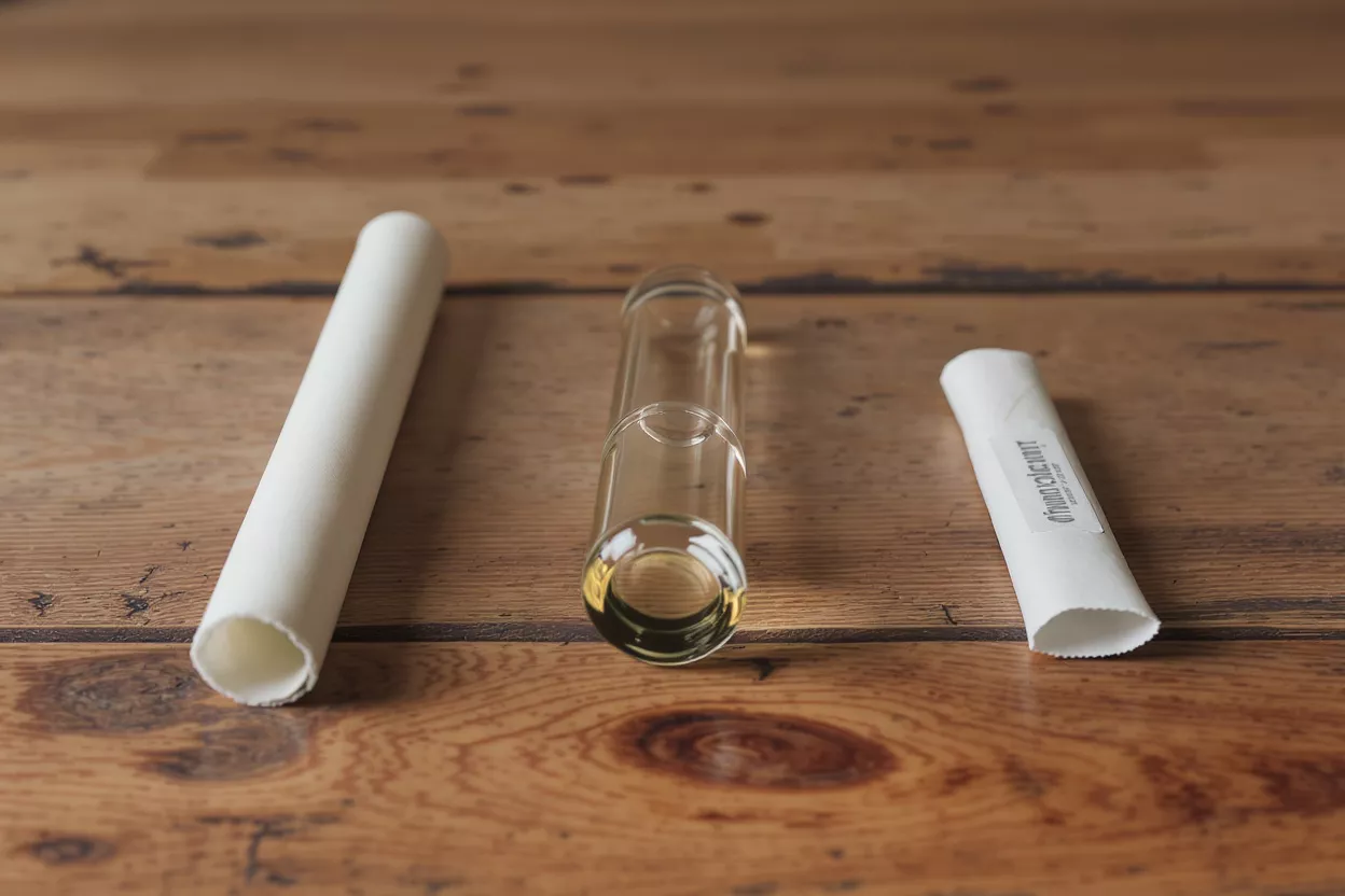 A glass vial containing yellow oil between two white paper sleeves on a wooden surface.