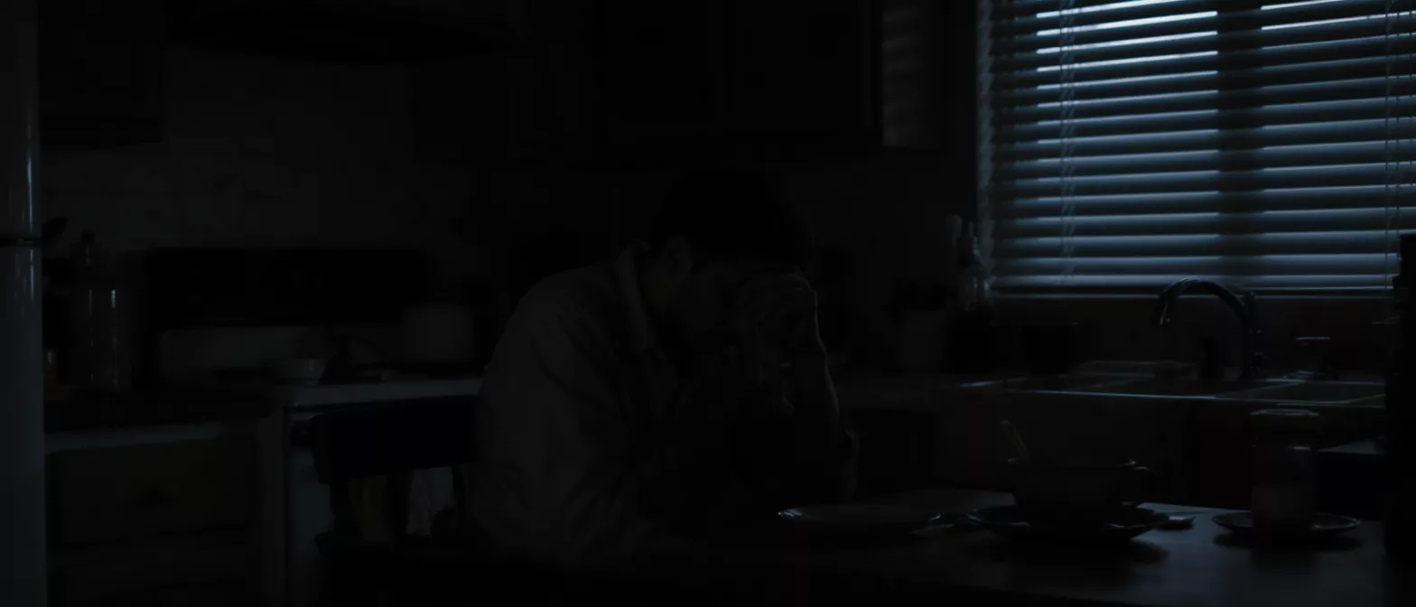 A man sits slumped at a table in a dark kitchen, illuminated by light from blinds.