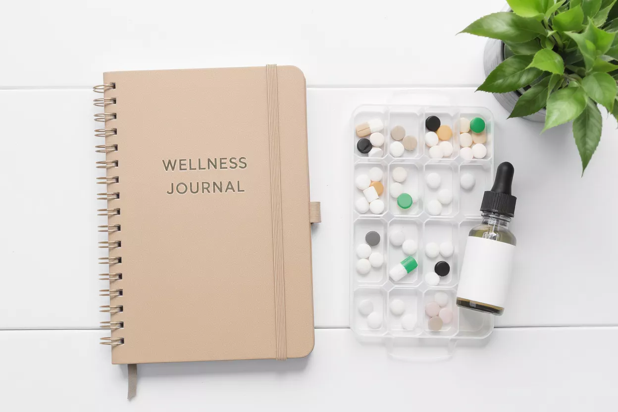 A beige wellness journal, pill organizer, dropper bottle, and plant on white surface.
