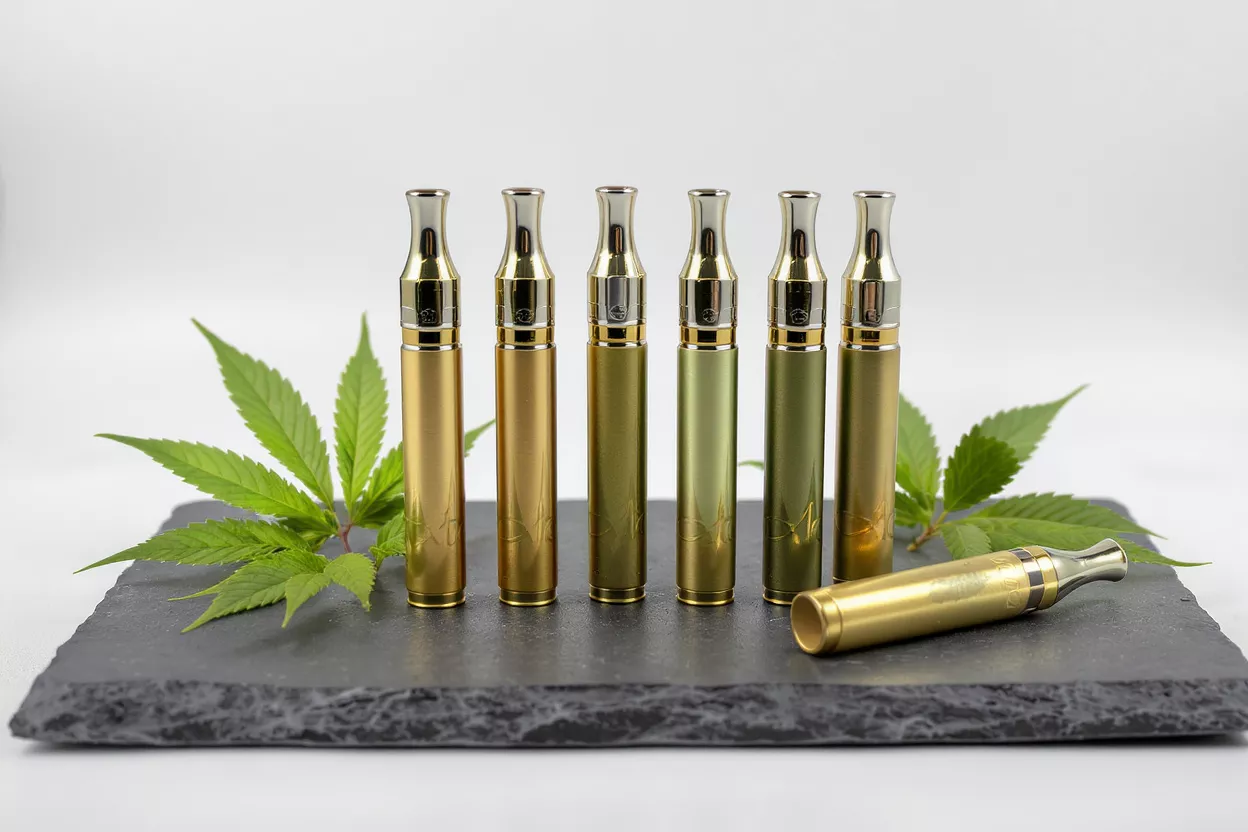 Six gold and green vape pens with cannabis leaves on a dark slate surface.